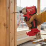 Milwaukee M18 Fuel brushless impact driver in action on a construction site, driving screws into wood framing with a worker's hand visible.